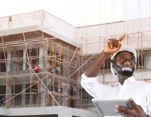 An smiling architect pointing to the sky and holding a grey colored tablet at a construction site.