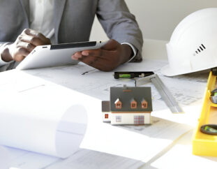 Building consultant with a tablet in hand, sitting by a table with floor plans, a white helmet, a building model, a ruler and level on top.